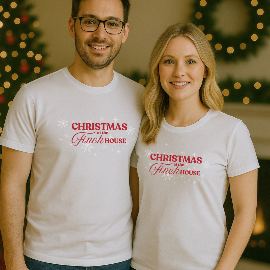 White T-shirt with “Christmas at the Finch House” design in red script, styled flat with festive decorations.
