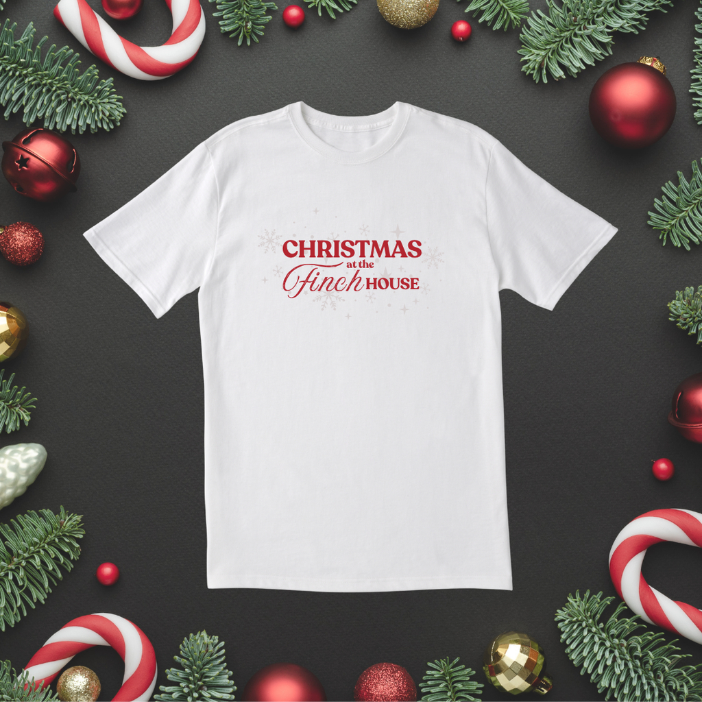White T-shirt with “Christmas at the Finch House” design in red script, styled flat with festive decorations.