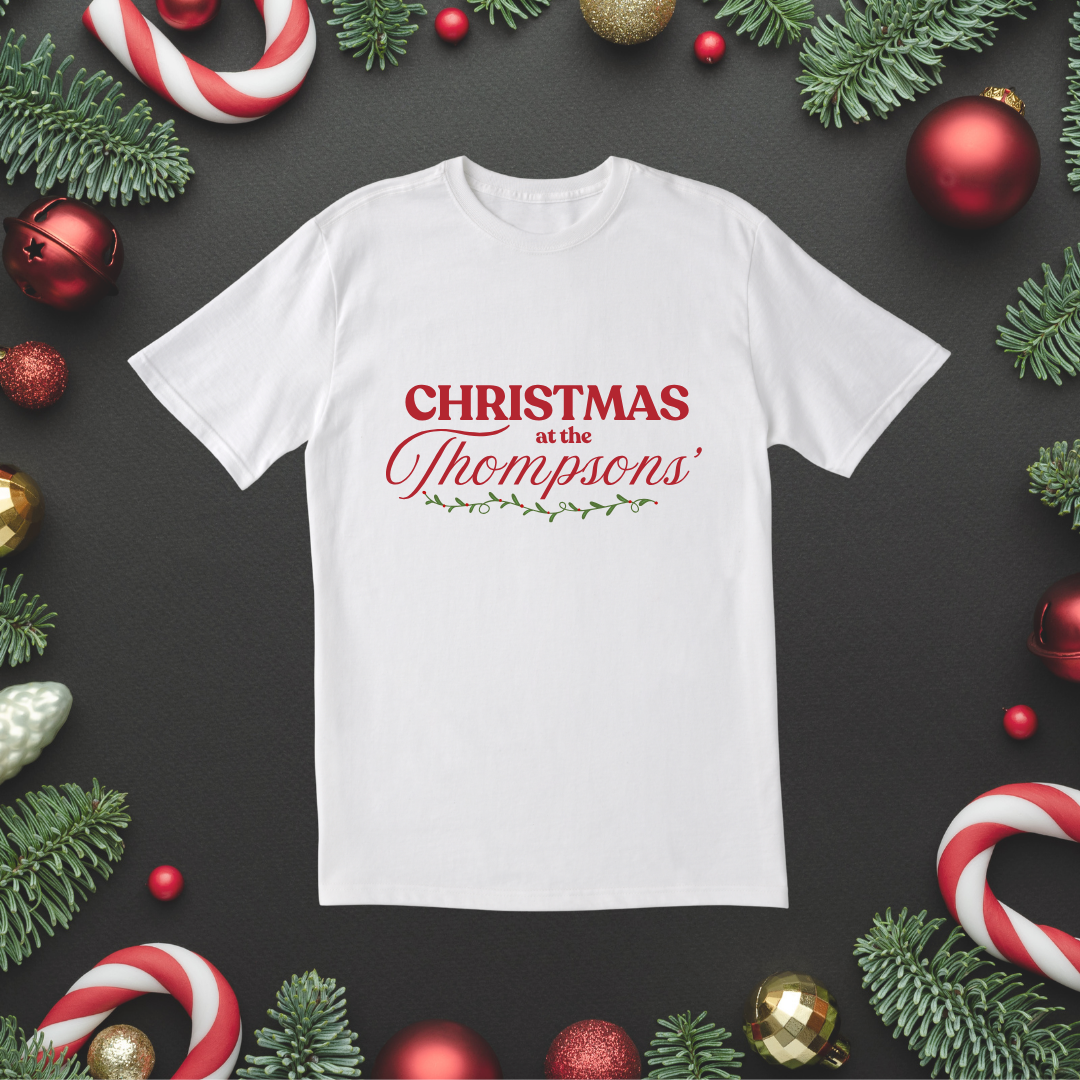 Woman wearing a white “Christmas at the Thompsons’” T-shirt in front of a Christmas tree and wreath.